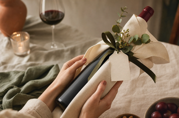 Hands presenting a linen-wrapped bottle of wine with ribbon and eucalyptus as a gift