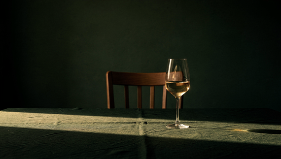 A single glass of white wine on a bare table in a quiet dim room, one empty chair behind